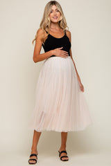woman wearing a black tank top and a light pink tulle maternity skirt, gently holding her baby bump