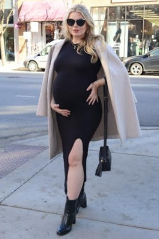 woman wearing a black maternity dress with a side slit and holding her baby bump, paired with a cream coat and sunglasses