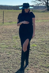 woman in black slit dress, teal wide-brim hat, and cowboy boots standing in a grassy field