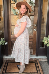 woman wearing a floral maternity dress and holding her baby bump, standing by a front door with a hat and boots