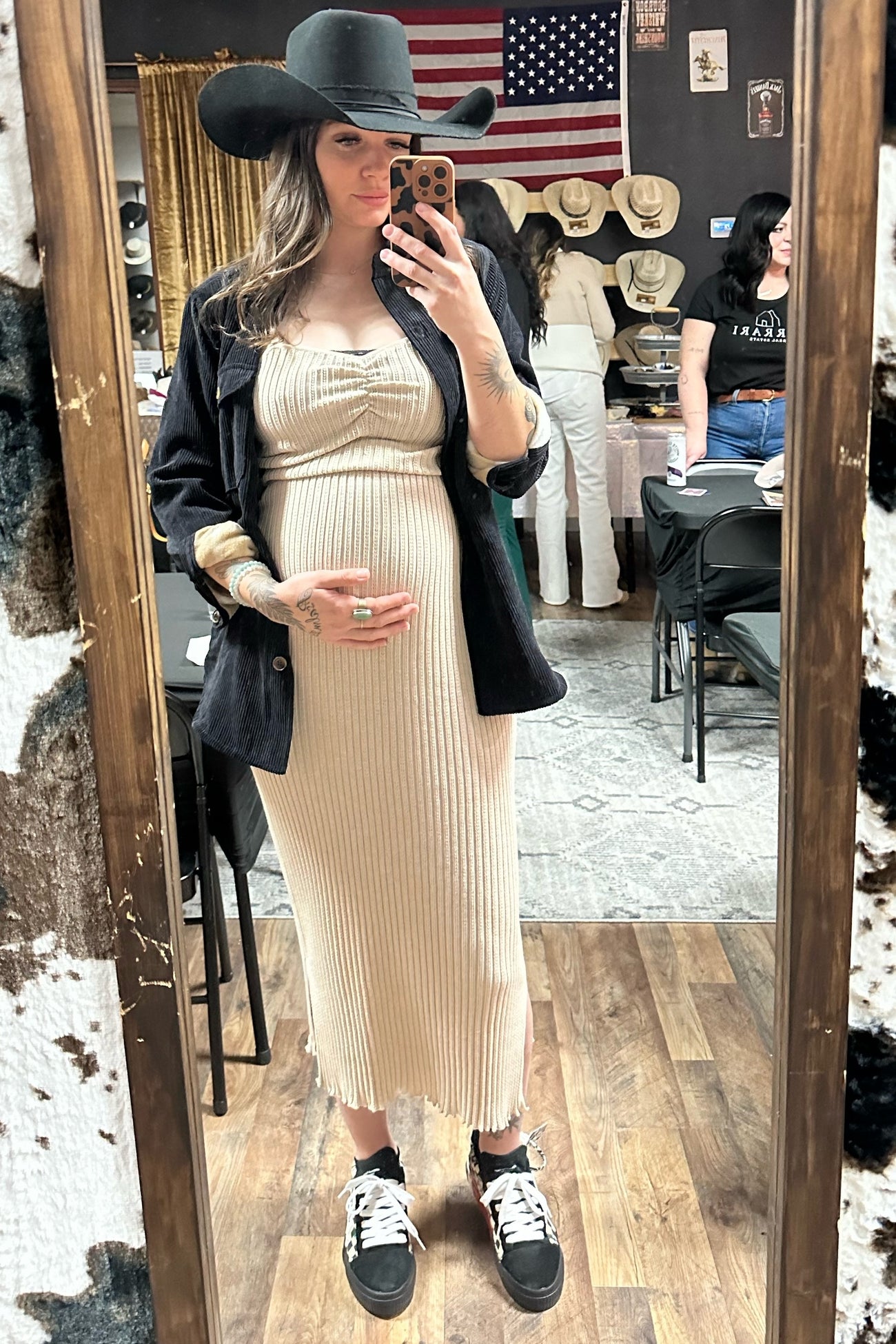 woman wearing a ribbed beige maternity dress with a black blazer and hat, holding her baby bump in a mirror selfie