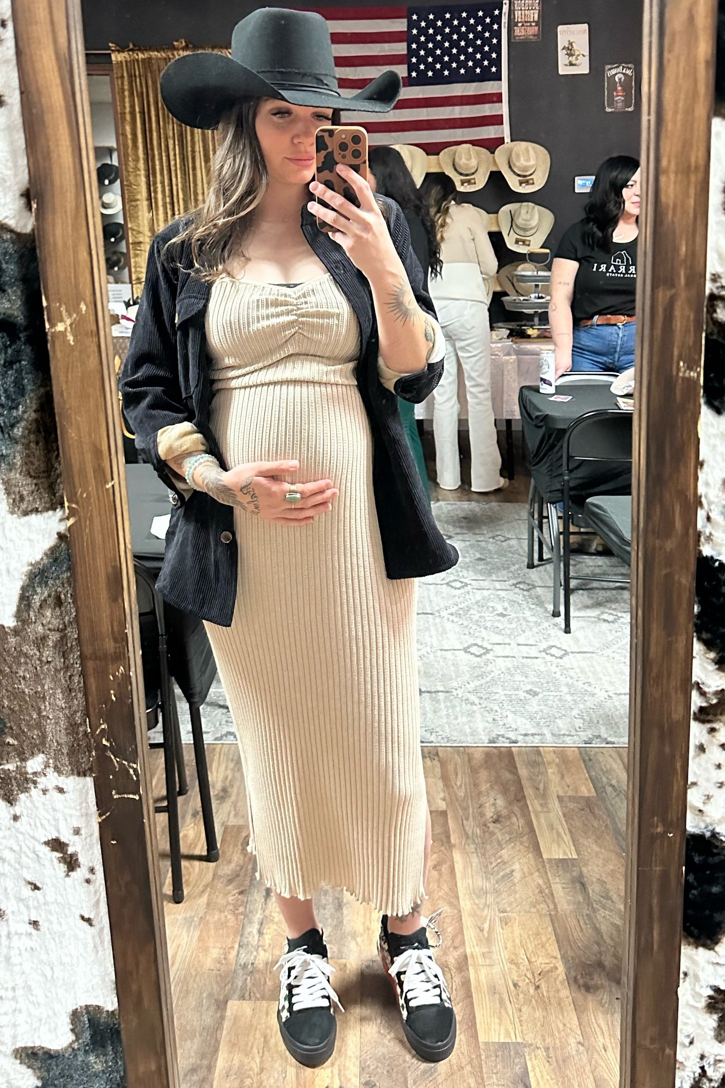woman wearing a ribbed beige maternity dress with a black blazer and hat, holding her baby bump in a mirror selfie