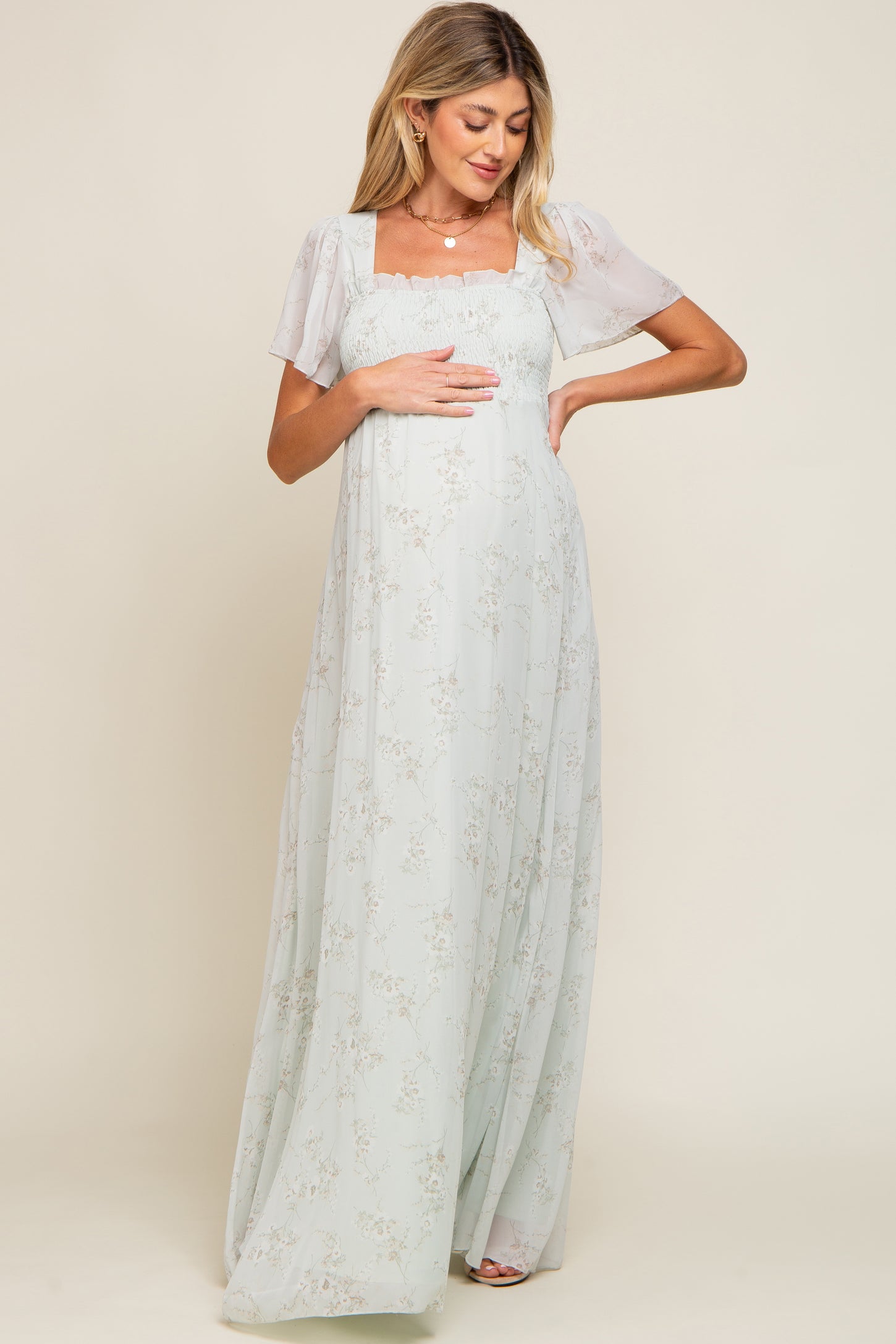 woman wearing a light green floral maternity dress with sheer short sleeves and a square neckline, holding her baby bump