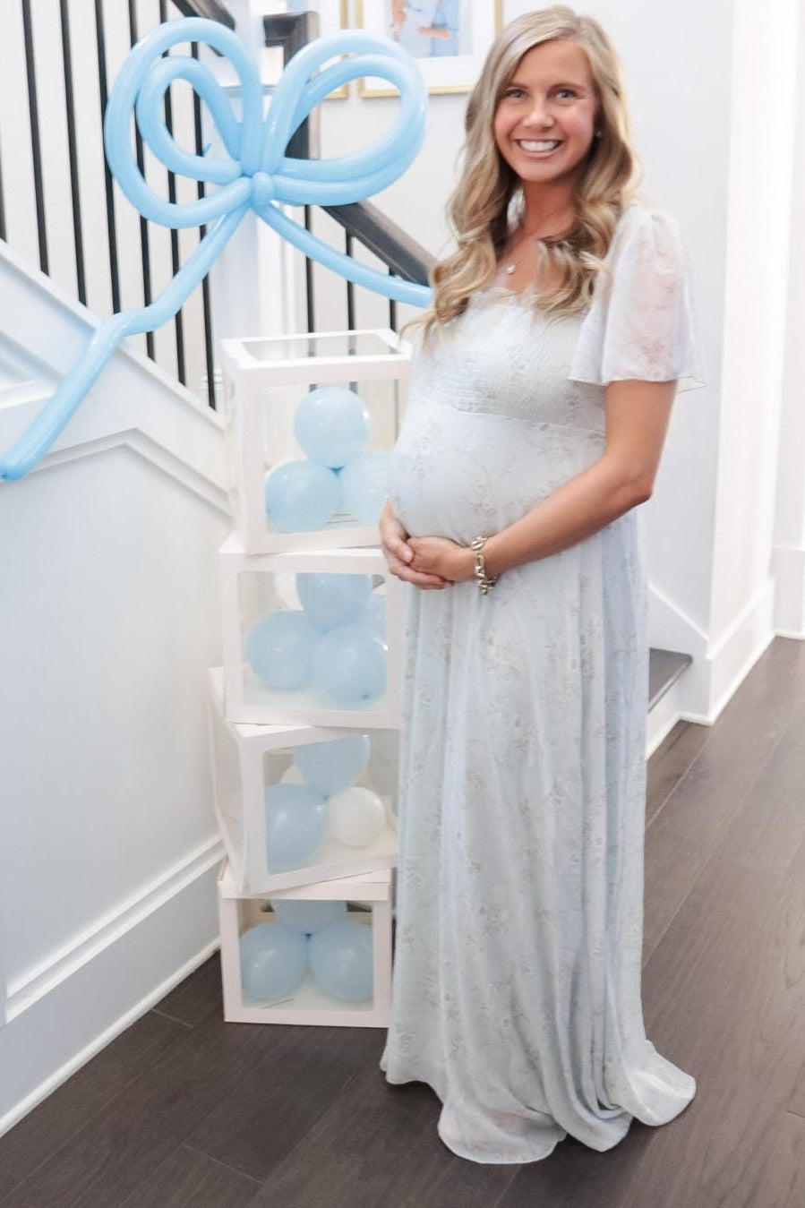 smiling pregnant woman in light blue floral maxi dress standing by blue balloon decorations at a baby shower