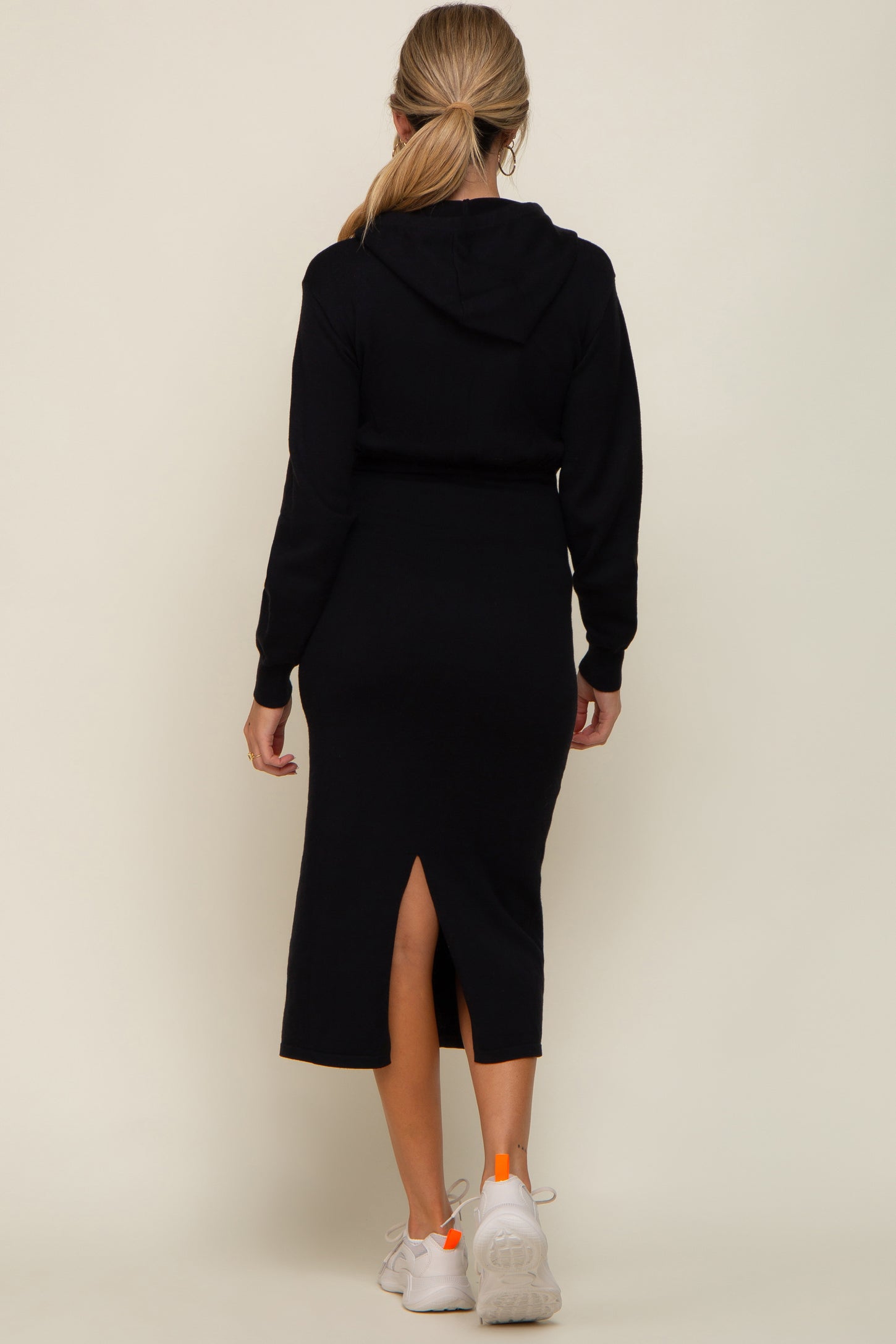 woman wearing a black maternity hoodie dress with a back slit, styled with white sneakers