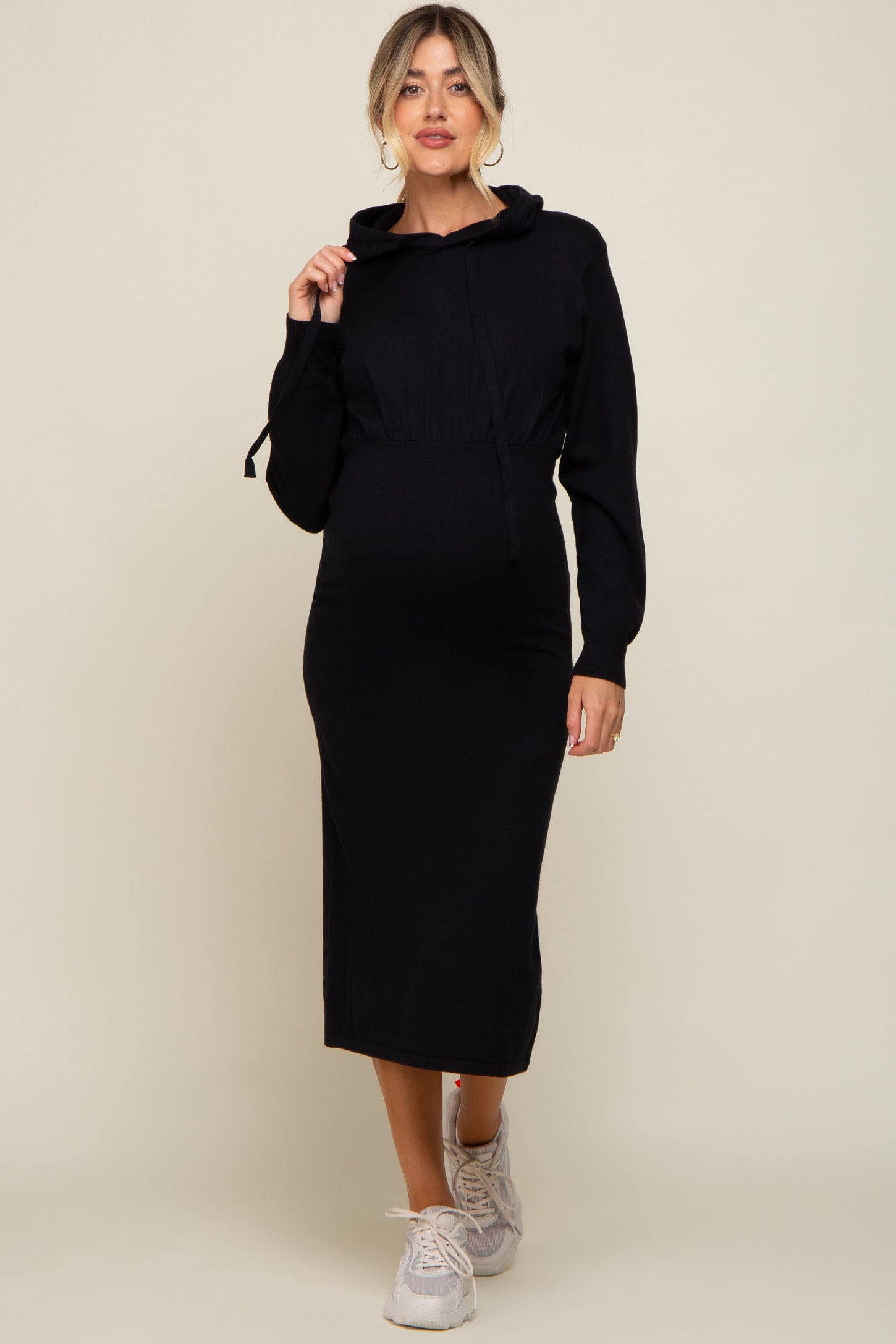 woman wearing a black maternity hoodie dress with a drawstring waist and long sleeves, holding the hood and showing her baby bump
