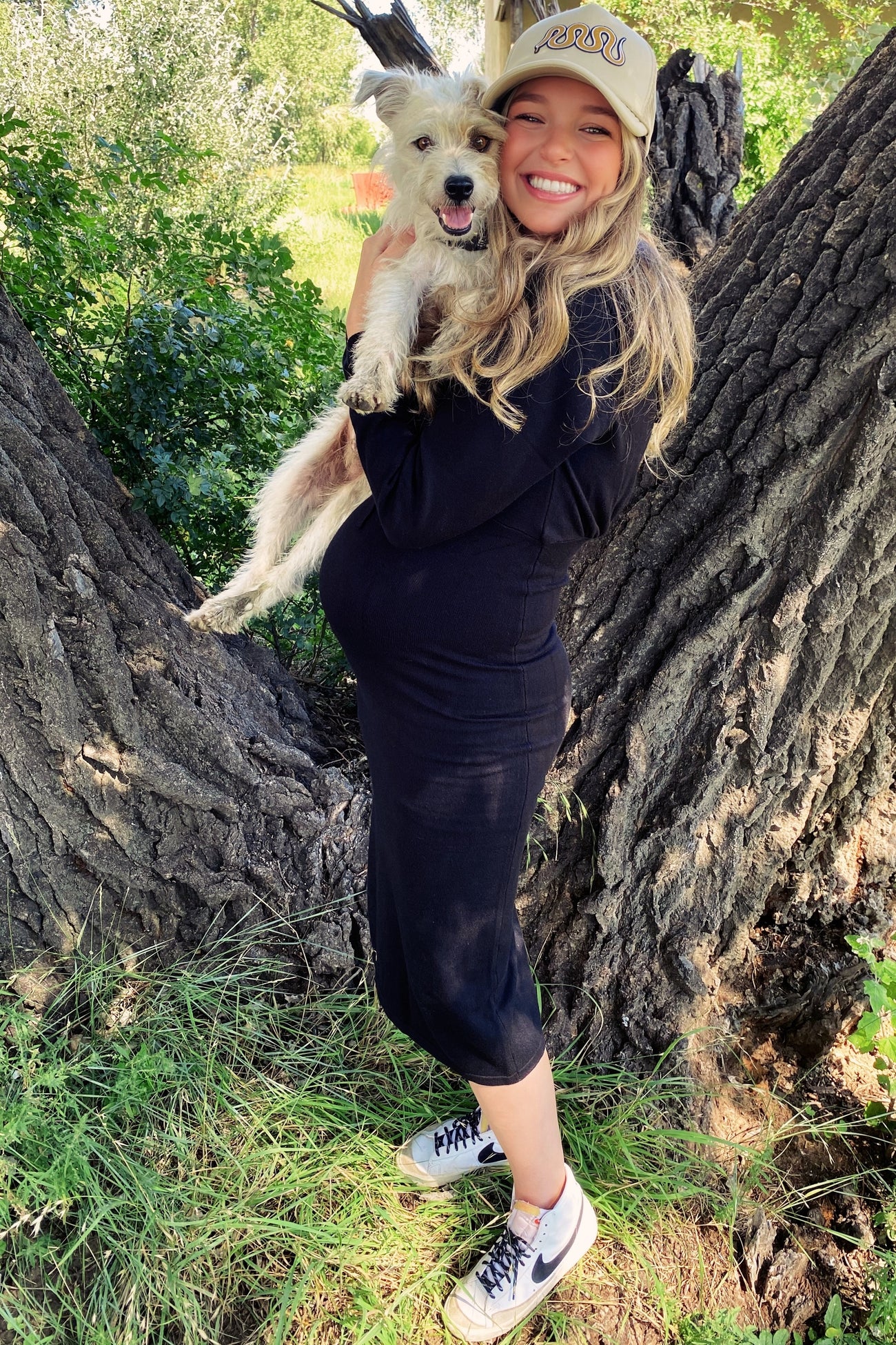 woman wearing a black maternity dress and holding a small dog, showing her baby bump against a natural background