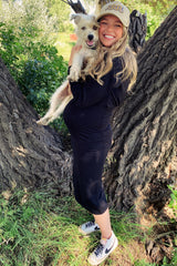 woman wearing a black maternity dress and holding a small dog, showing her baby bump against a natural background