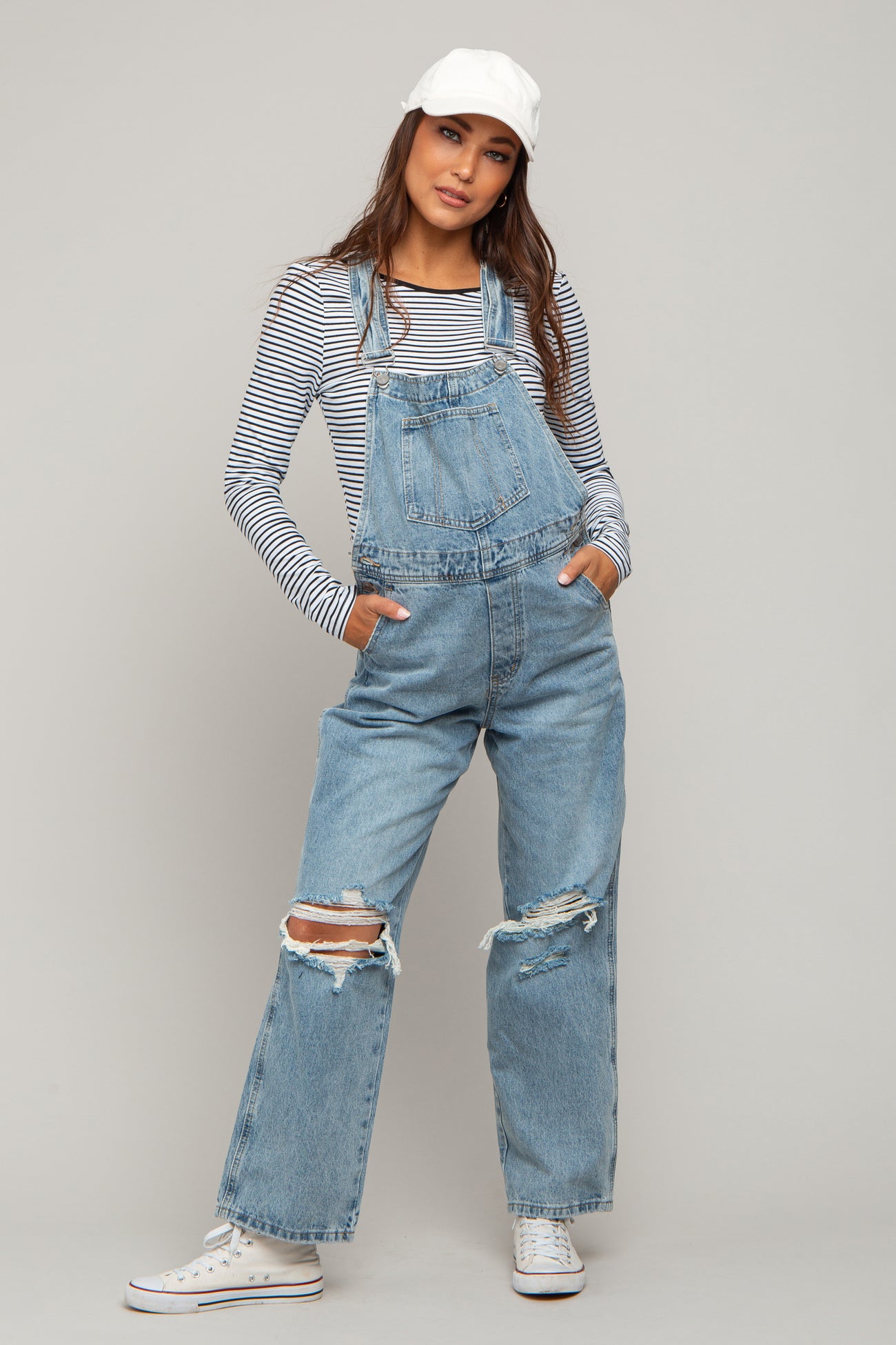 woman wearing a striped long sleeve maternity top with distressed denim overalls and a white cap, hands in pockets