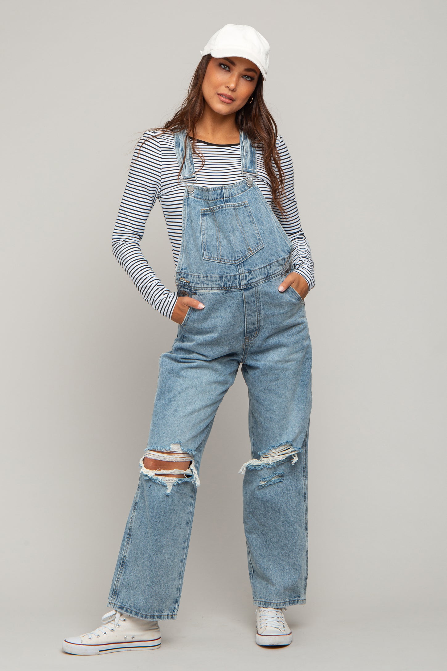 woman wearing a striped long sleeve maternity top with distressed denim overalls and a white cap, hands in pockets