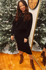 woman wearing a black long sleeve top and matching midi skirt, posing in front of a mirror with a festive backdrop