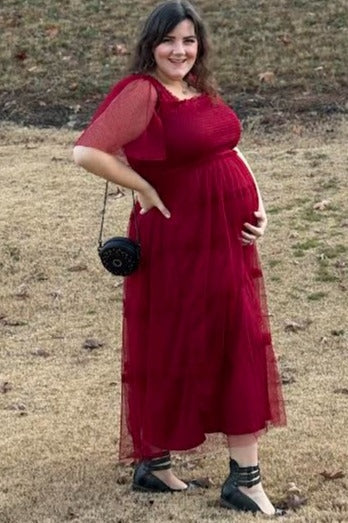 woman wearing a red maternity maxi dress with sheer sleeves and holding her baby bump outdoors