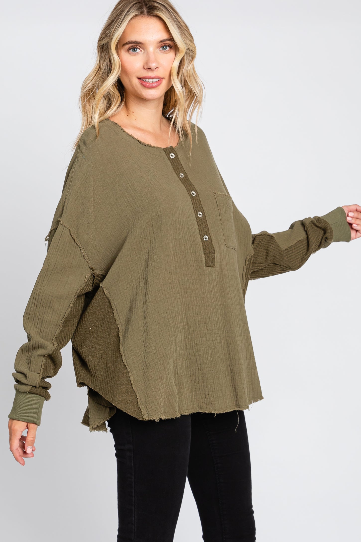 woman wearing an oversized olive green long sleeve button-up top with a pocket and raw hem, paired with black pants