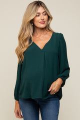 woman wearing a dark green maternity top with a v-neck and long sleeves, smiling and standing with hands on her belly