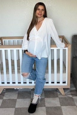 woman wearing a white blouse and distressed jeans, showcasing her baby bump while standing next to a baby crib