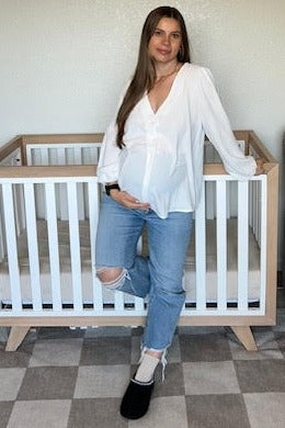 woman wearing a white button-up maternity top and distressed blue jeans, showing her baby bump next to a crib