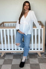 woman wearing a white button-up maternity top and distressed blue jeans, showing her baby bump next to a crib