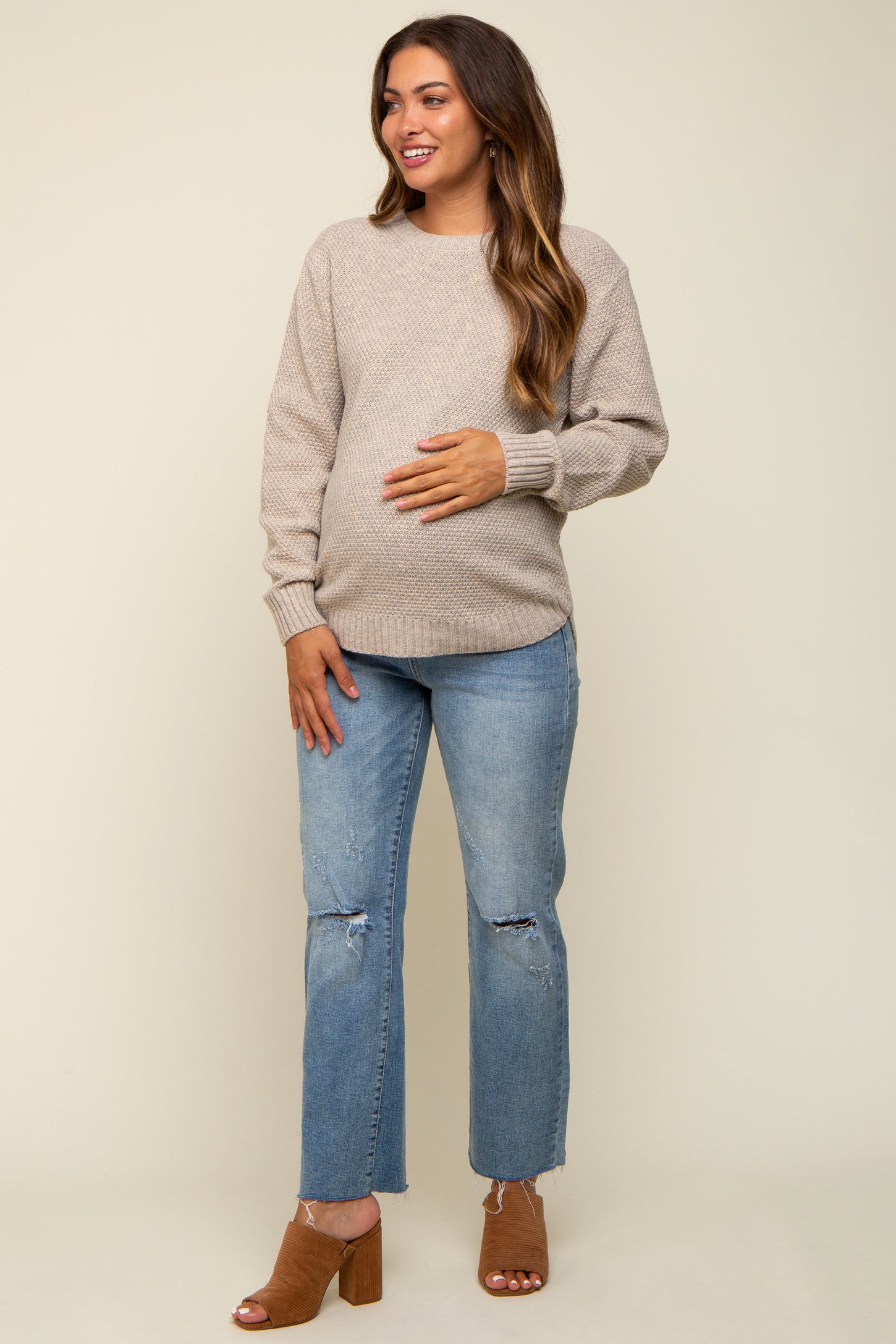 woman wearing a light beige maternity sweater and distressed blue jeans, showing her baby bump, smiling at the camera