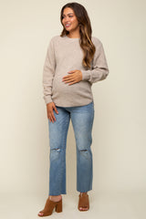 woman wearing a light beige maternity sweater and distressed blue jeans, showing her baby bump, smiling at the camera