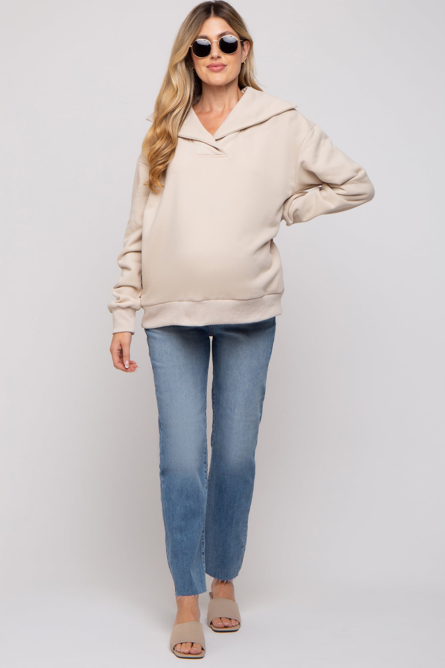 woman wearing a beige maternity hoodie with a loose fit and long sleeves, paired with light blue jeans and sunglasses, showcasing a baby bump