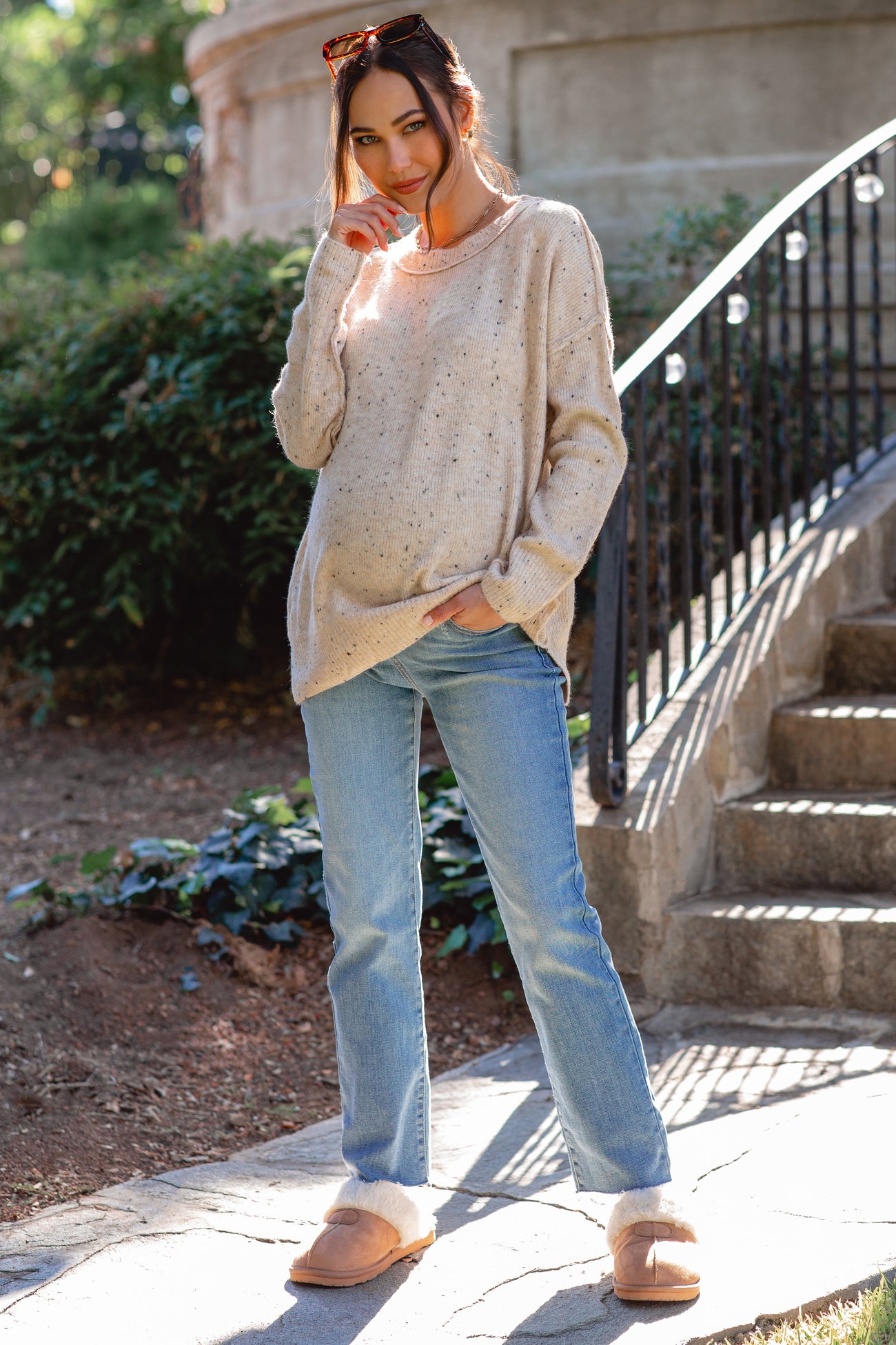 woman wearing a light beige maternity sweater and light blue maternity jeans, showing her baby bump, standing outdoors