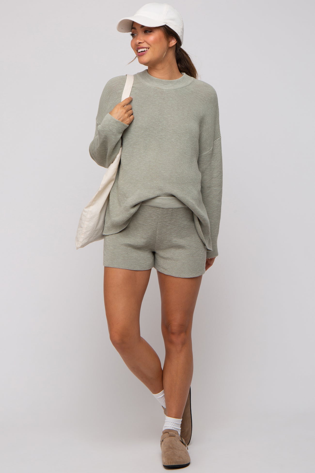 woman wearing a light green maternity sweater and shorts set with a cap and carrying a tote bag