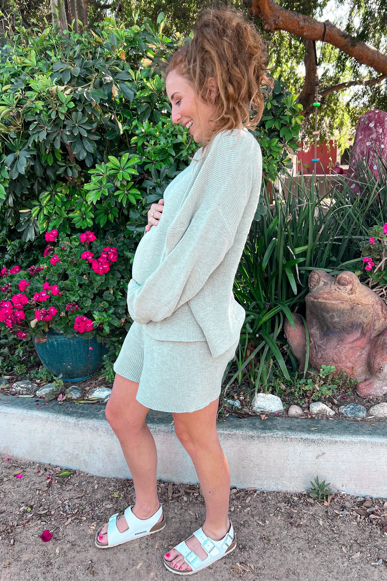 woman wearing a light gray maternity cardigan and matching shorts, cradling her baby bump in a garden setting