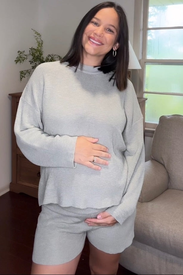 woman wearing a light grey maternity sweater and shorts, holding her baby bump, smiling in a living room setting
