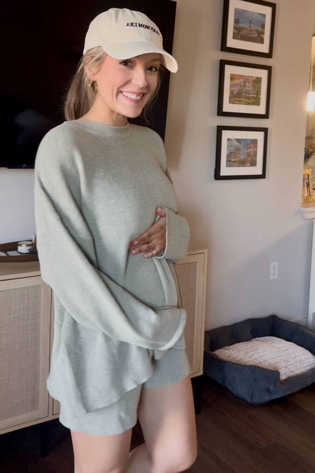 woman wearing a light green oversized maternity sweater and shorts, holding her baby bump, smiling at the camera