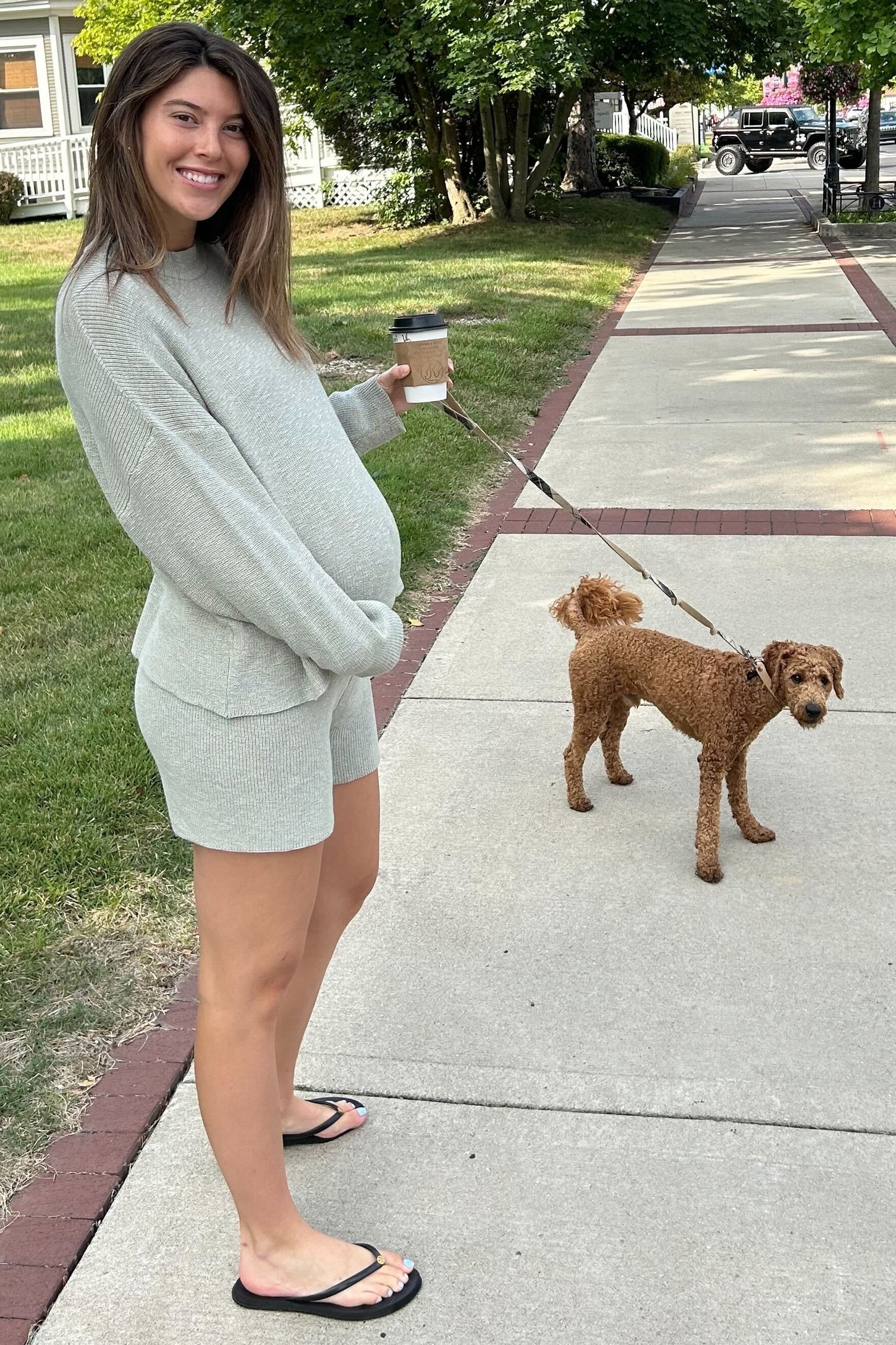 woman wearing a light gray maternity sweater and shorts, holding a coffee cup and standing beside a brown dog