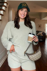 woman wearing a light green oversized sweater and matching shorts, holding a coffee cup, with a baseball cap and handbag