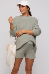 woman wearing a light green oversized knit maternity sweater and matching shorts, holding a tote bag and wearing a cap