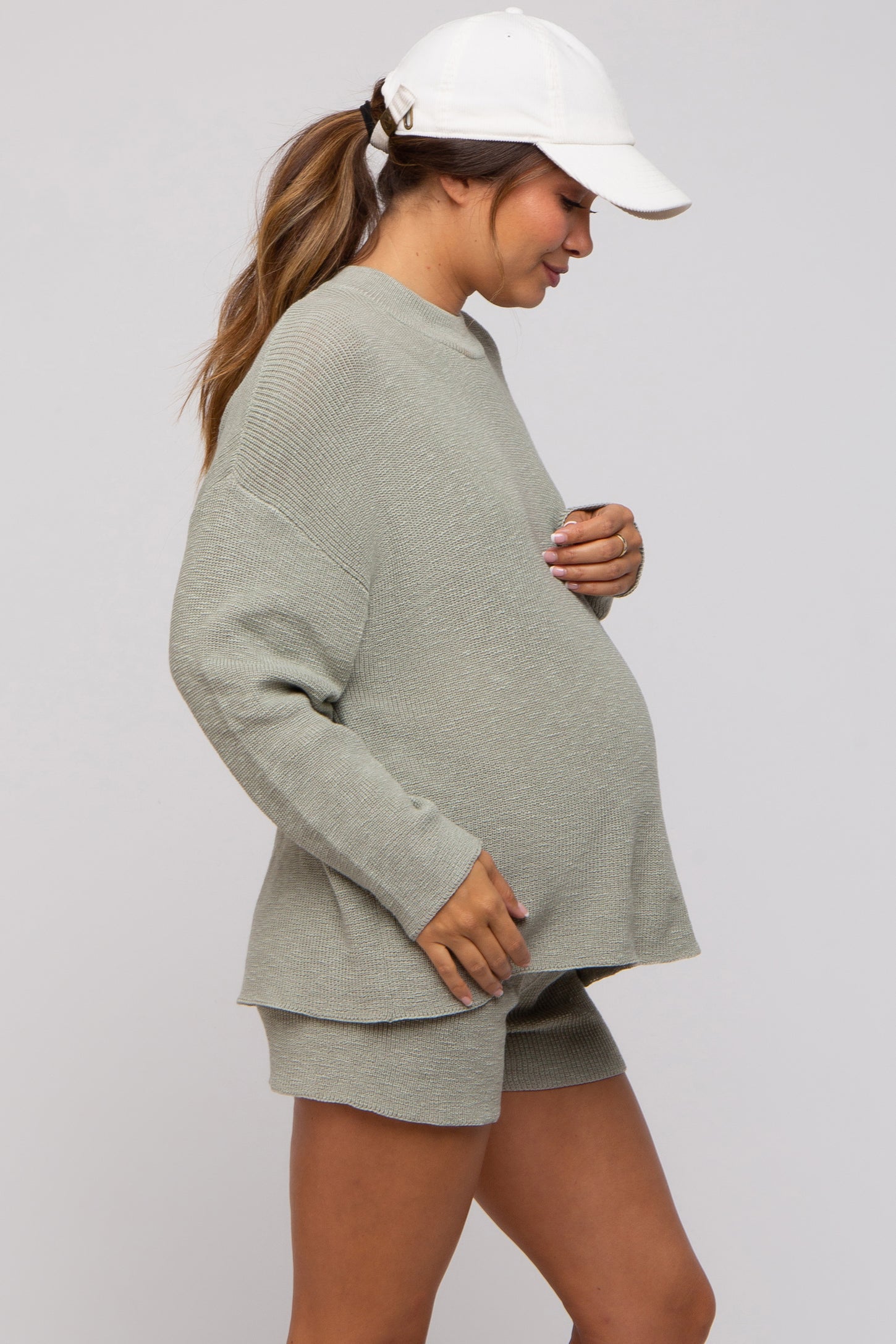 woman wearing a light green maternity sweatshirt and shorts, gently cradling her baby bump