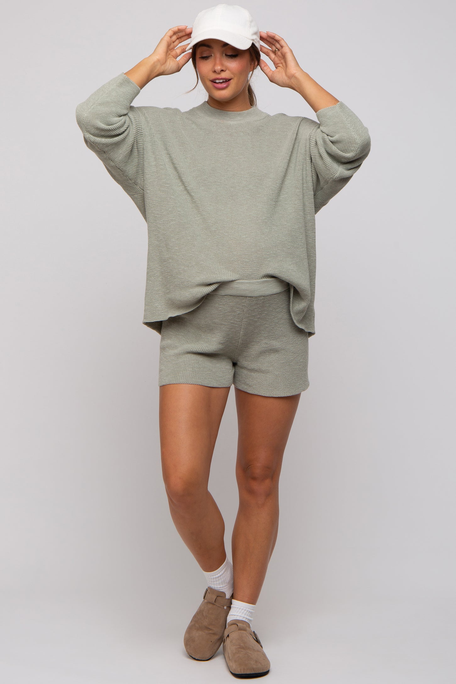woman wearing a light green maternity sweater and matching shorts, styled with a white cap and light brown shoes