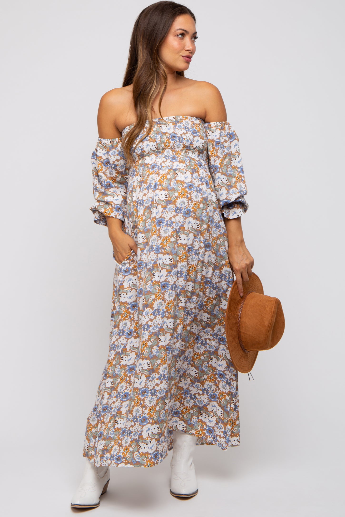 woman wearing a floral off-the-shoulder maternity maxi dress and holding a brown hat, showcasing her baby bump