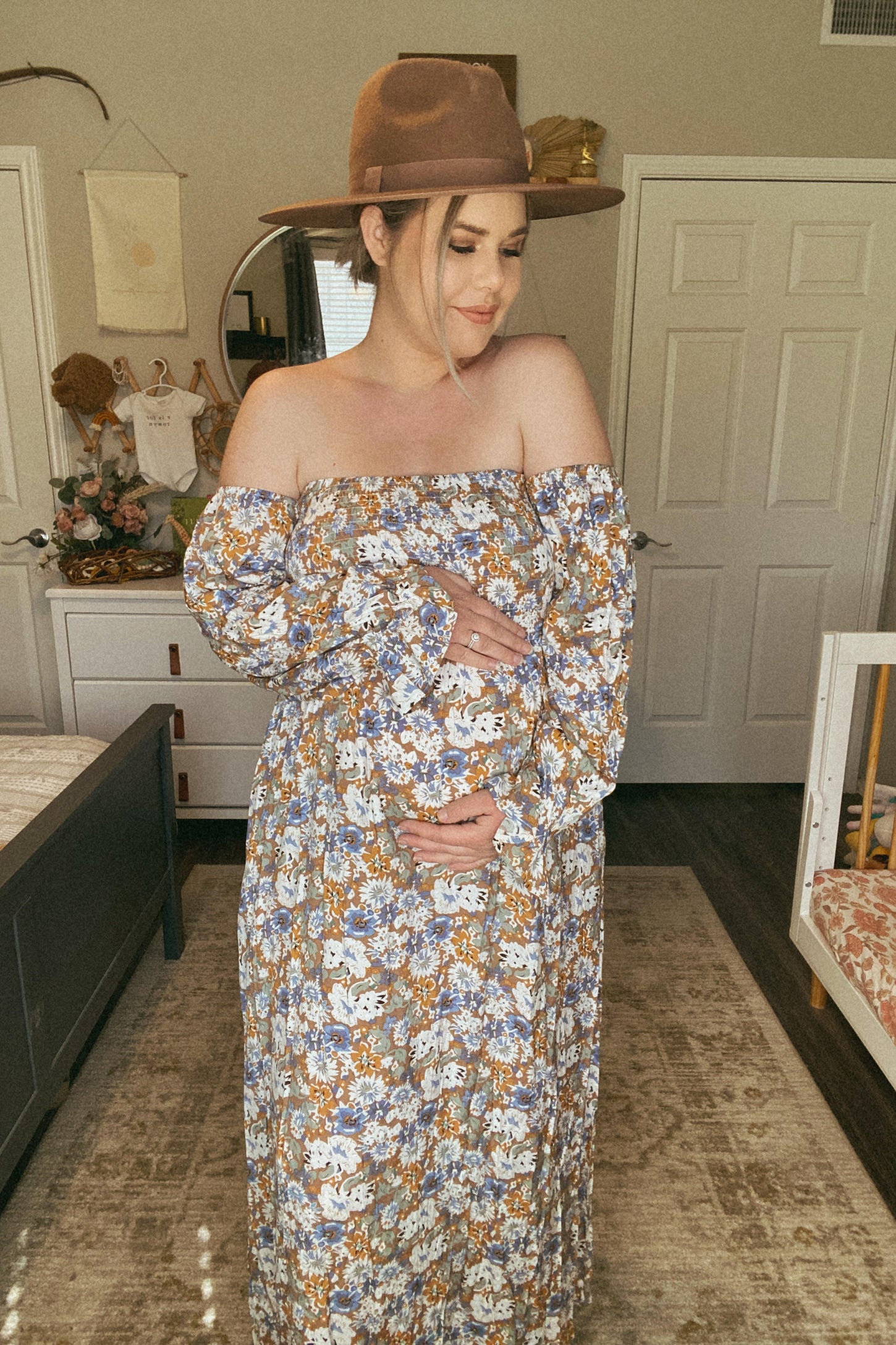 woman wearing a floral off-shoulder maternity dress and holding her baby bump, accessorized with a wide-brim hat