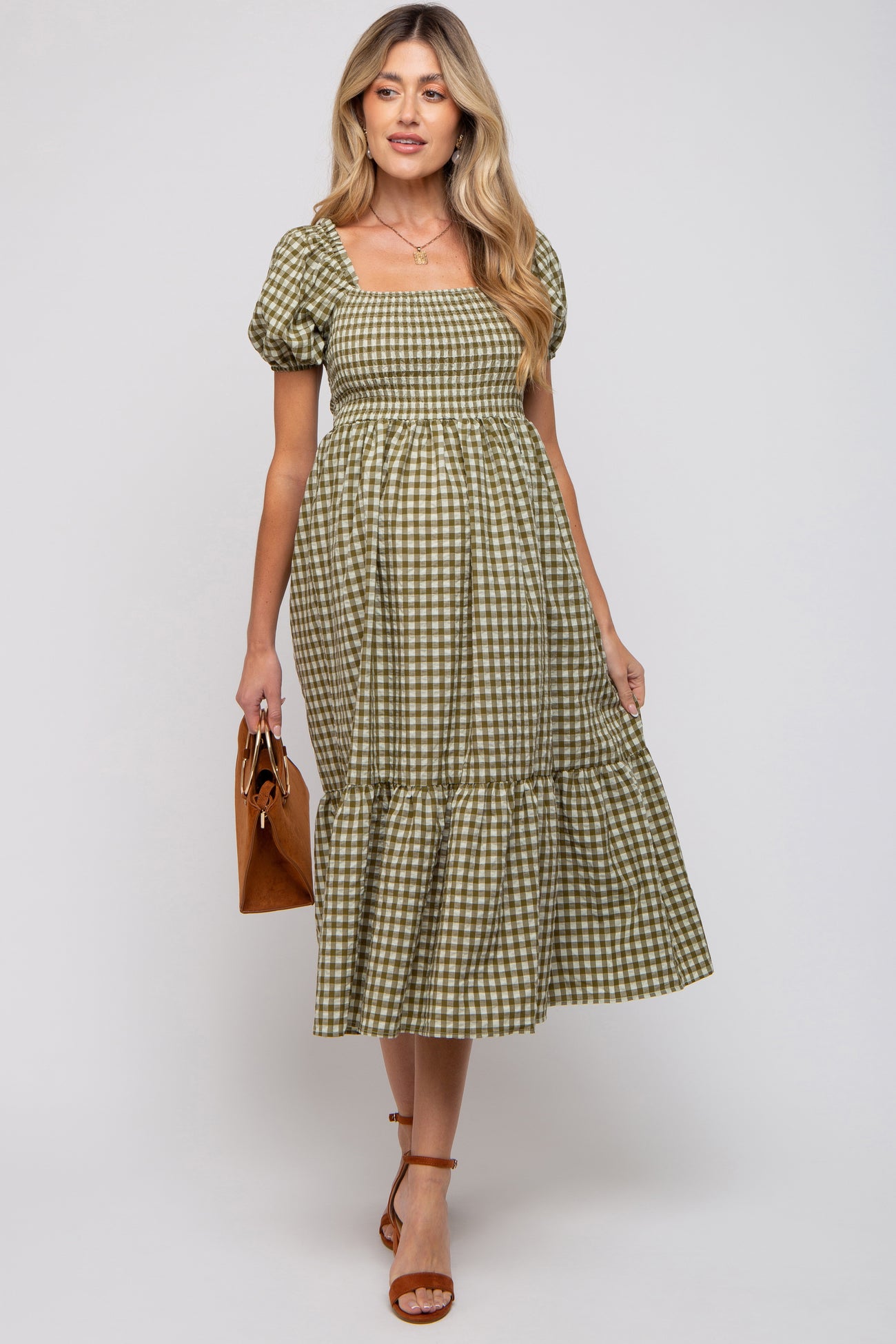 woman wearing a green and white checkered maternity dress with puffed sleeves and a tiered hem, holding a brown purse