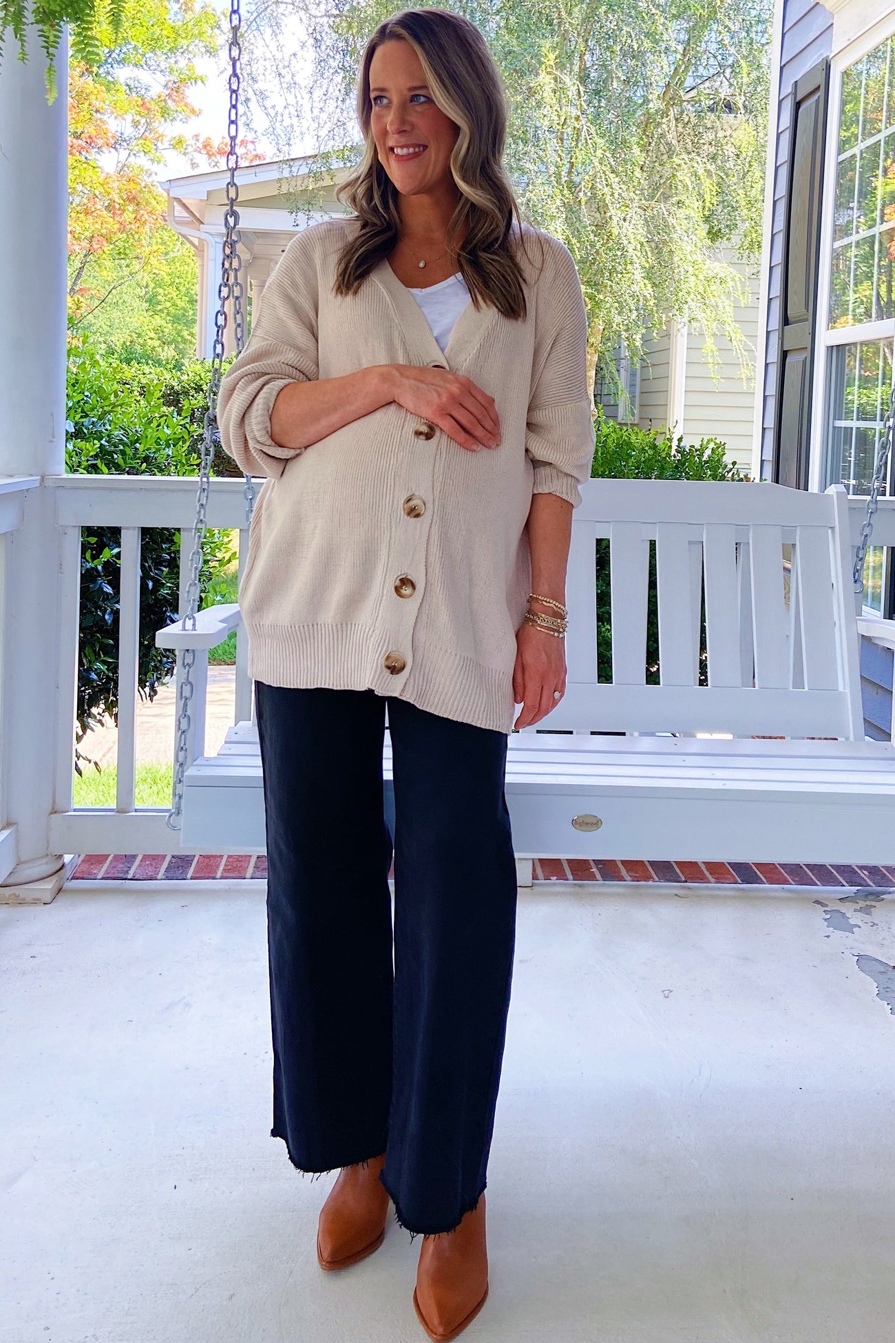 woman wearing a beige cardigan and a white tank top paired with black wide-leg pants, standing by a porch swing