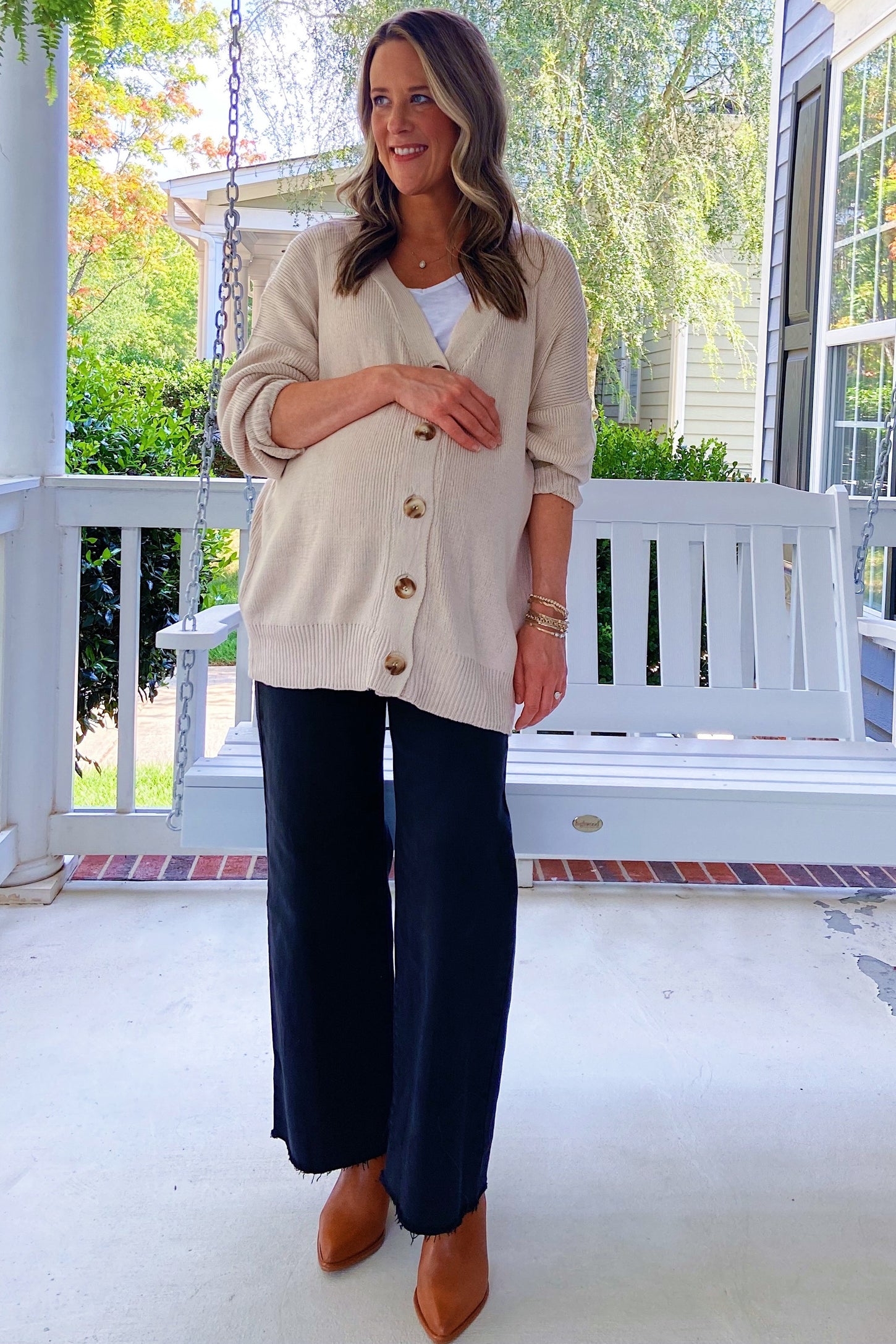 woman wearing a beige cardigan and a white tank top paired with black wide-leg pants, standing by a porch swing