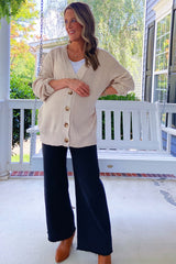 woman wearing a cream maternity cardigan with buttons, white tank top, dark wide-leg pants, and a visible baby bump