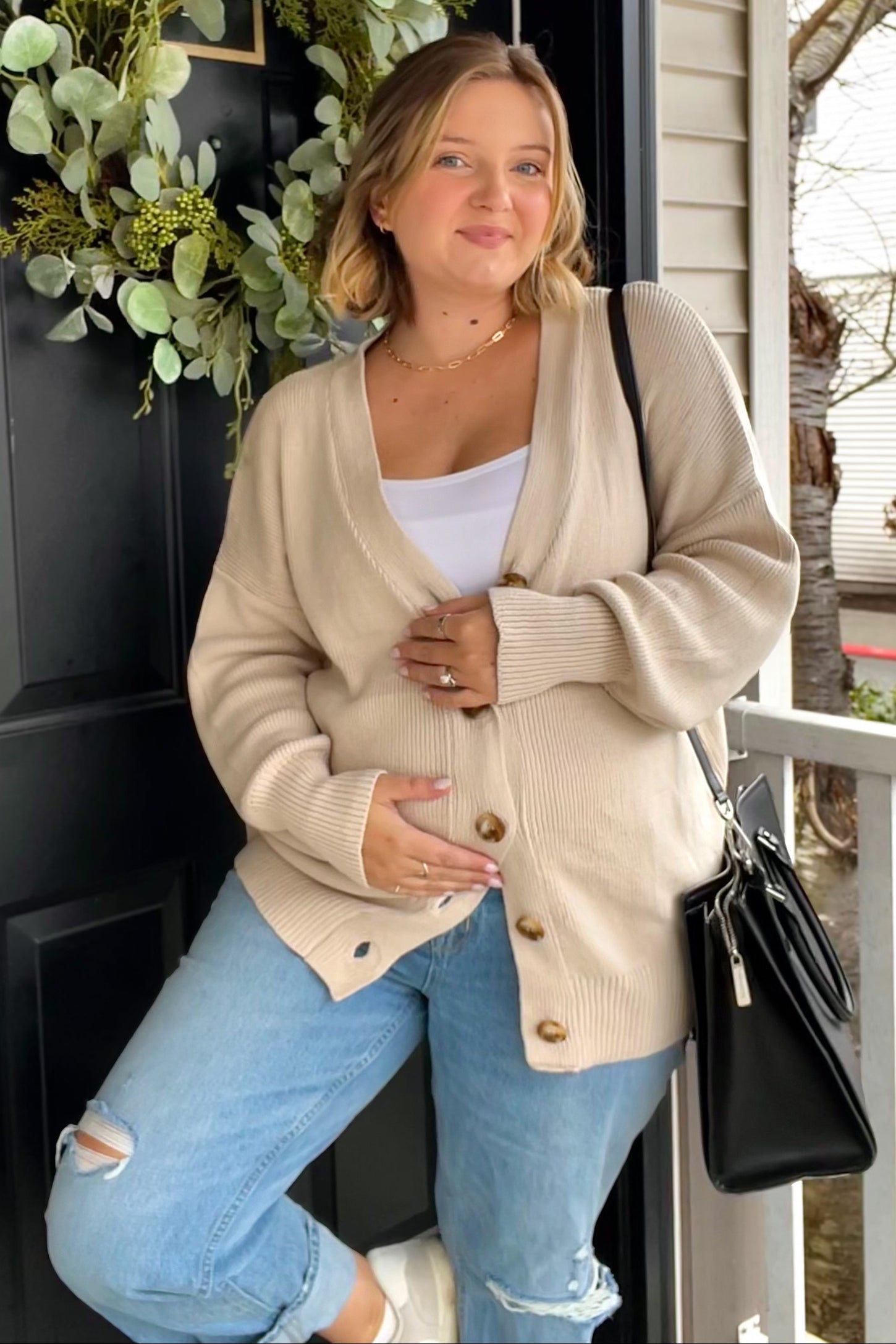 woman wearing a beige knit maternity cardigan, holding her baby bump, paired with light blue distressed jeans and a black handbag