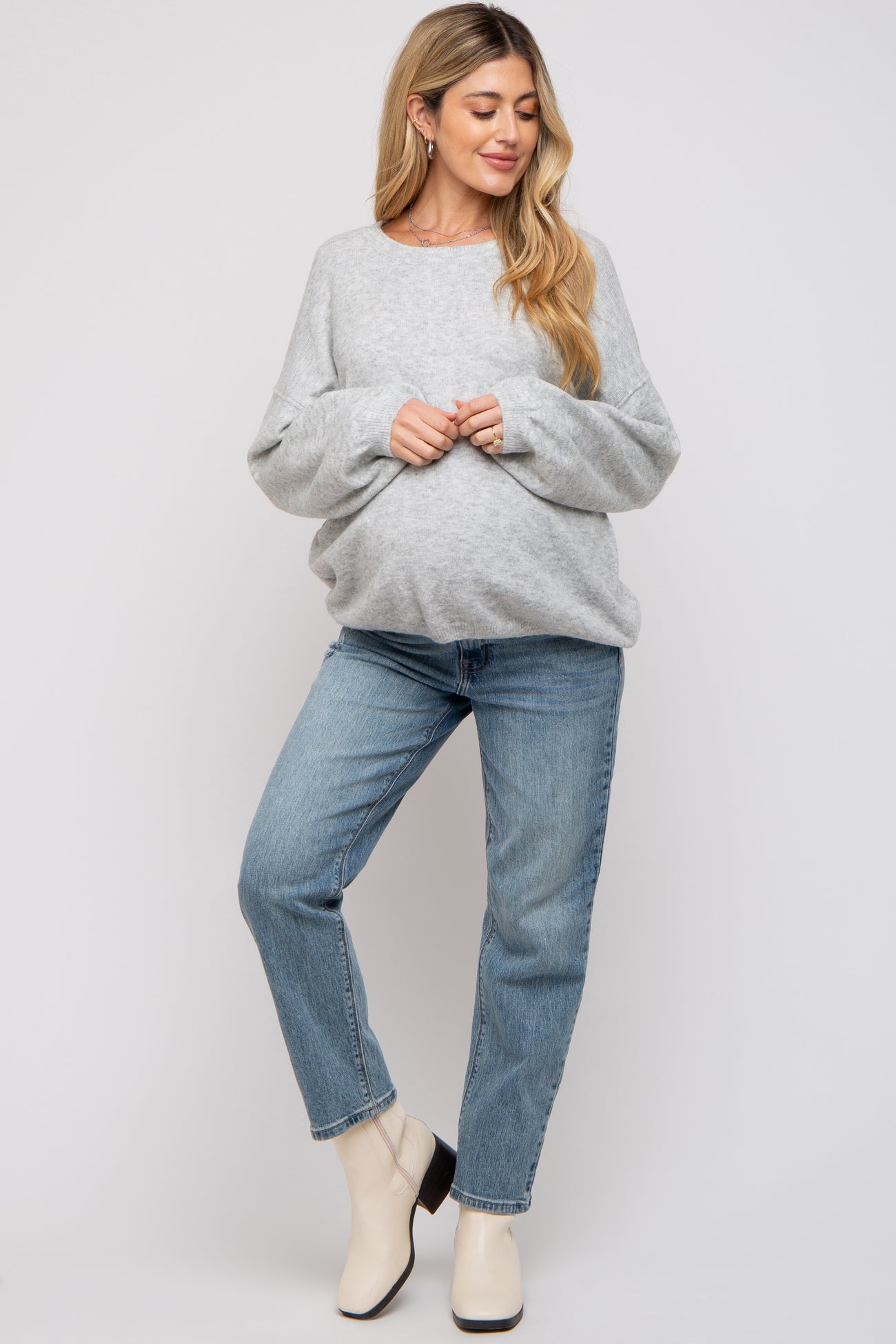 woman wearing a gray maternity sweater and blue jeans, gently cradling her baby bump with a relaxed pose