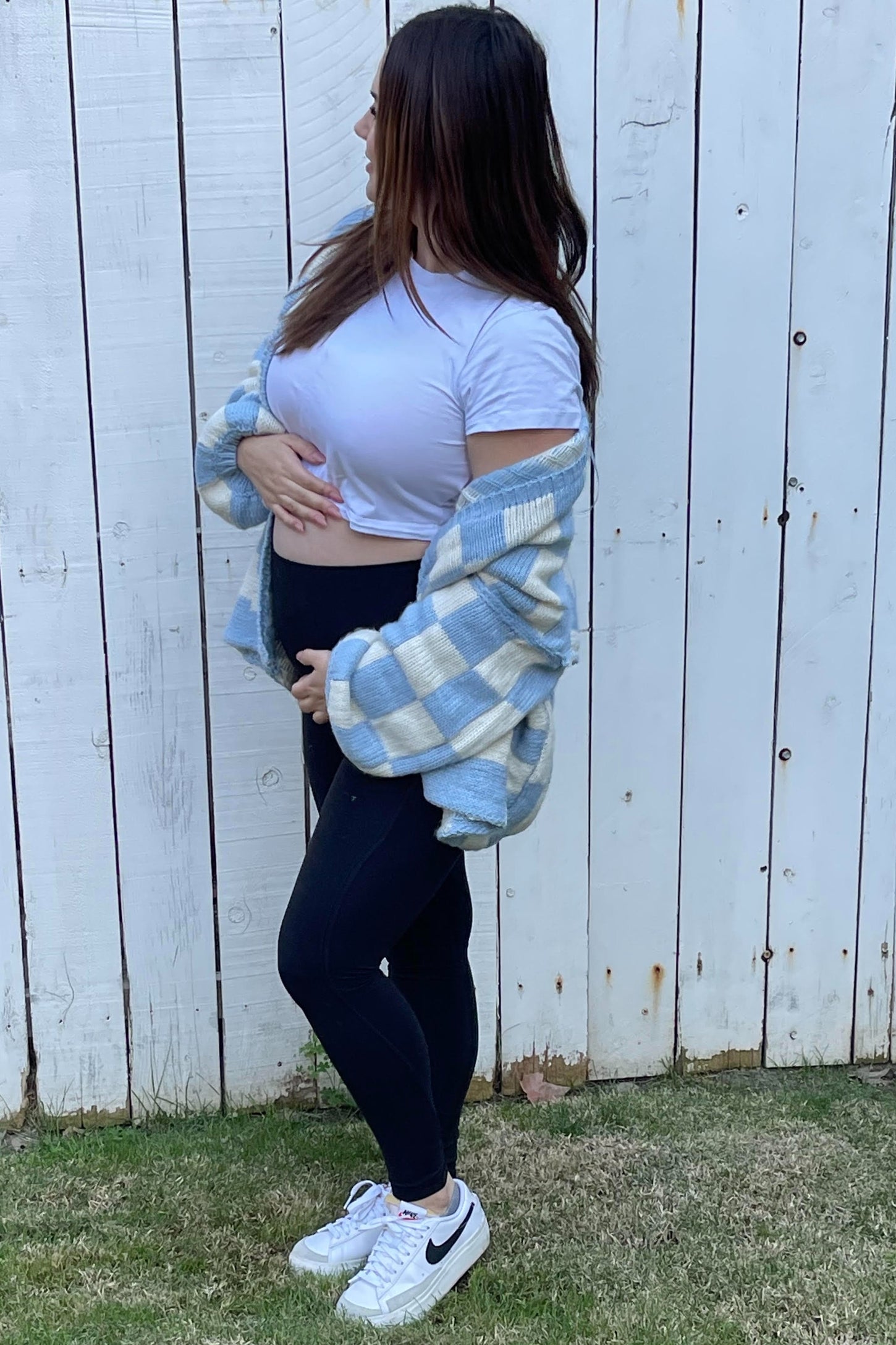 woman wearing a white short sleeve top and black leggings, holding her baby bump, layered with a blue plaid oversized shirt