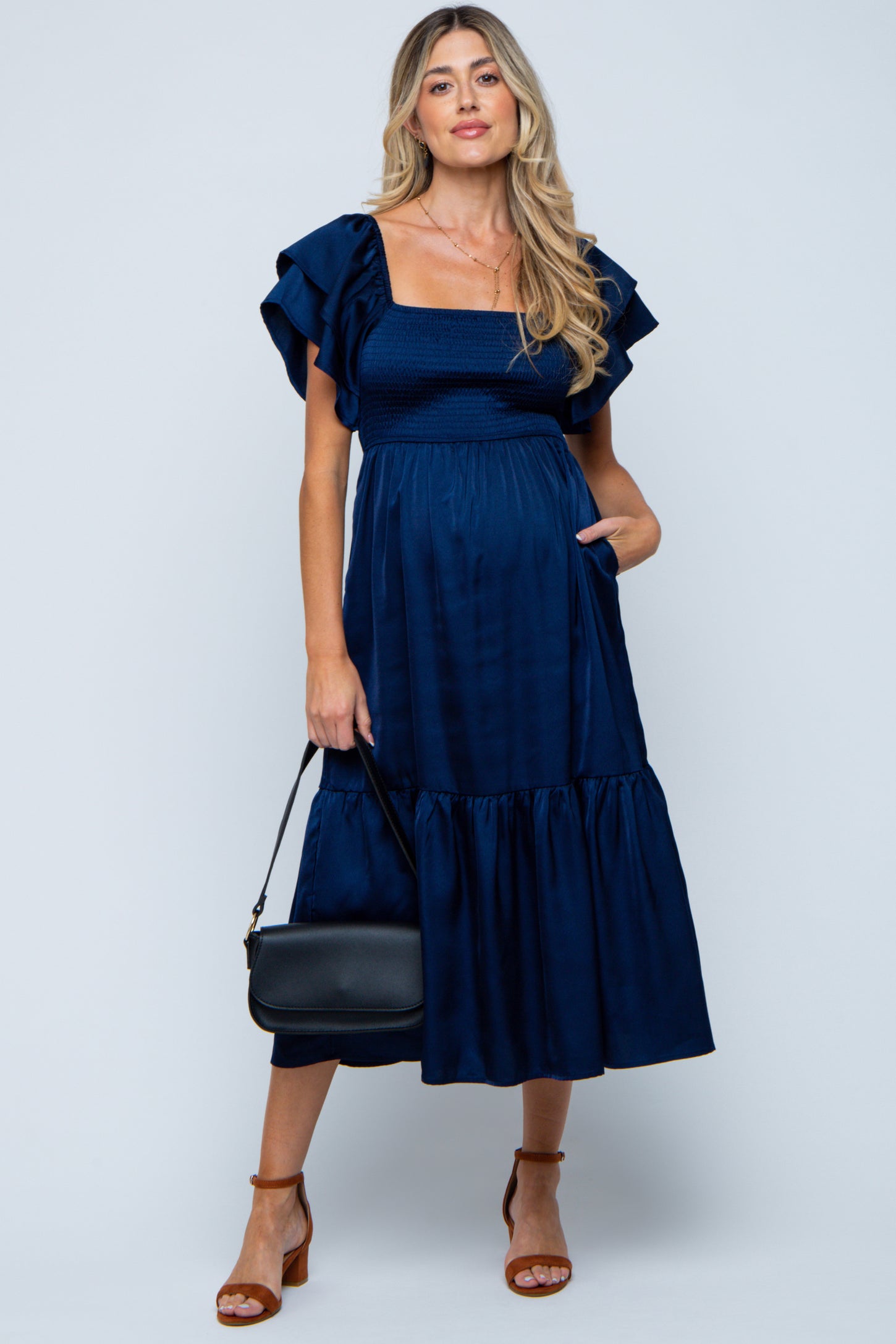 woman wearing a navy blue maternity dress with ruffled sleeves, fitted bodice, and tiered skirt, holding a black handbag
