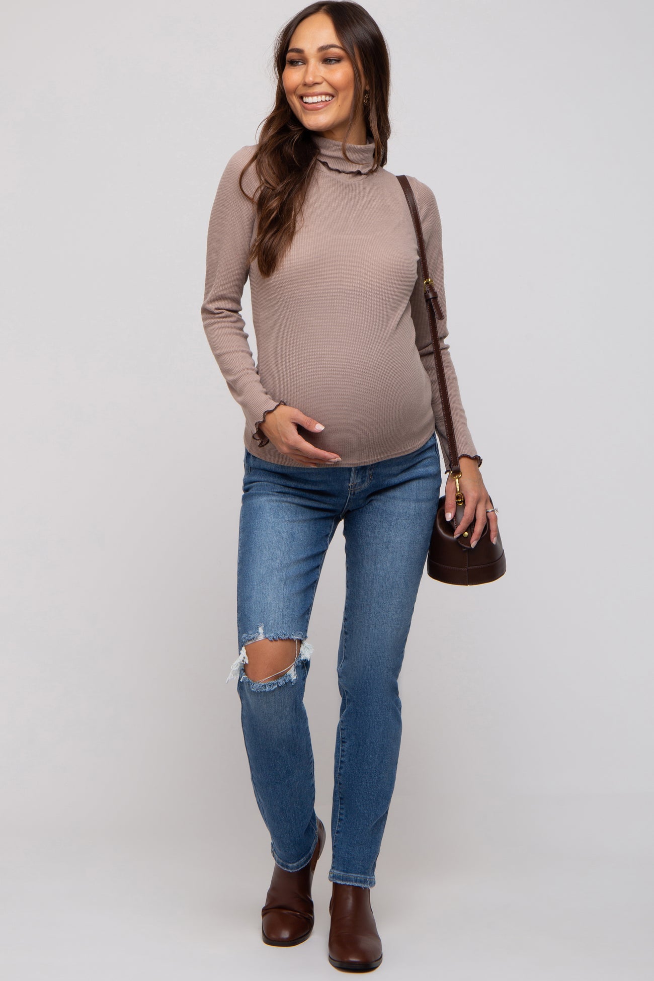 woman wearing a taupe long sleeve maternity top and distressed blue jeans, showing her baby bump with a brown handbag.
