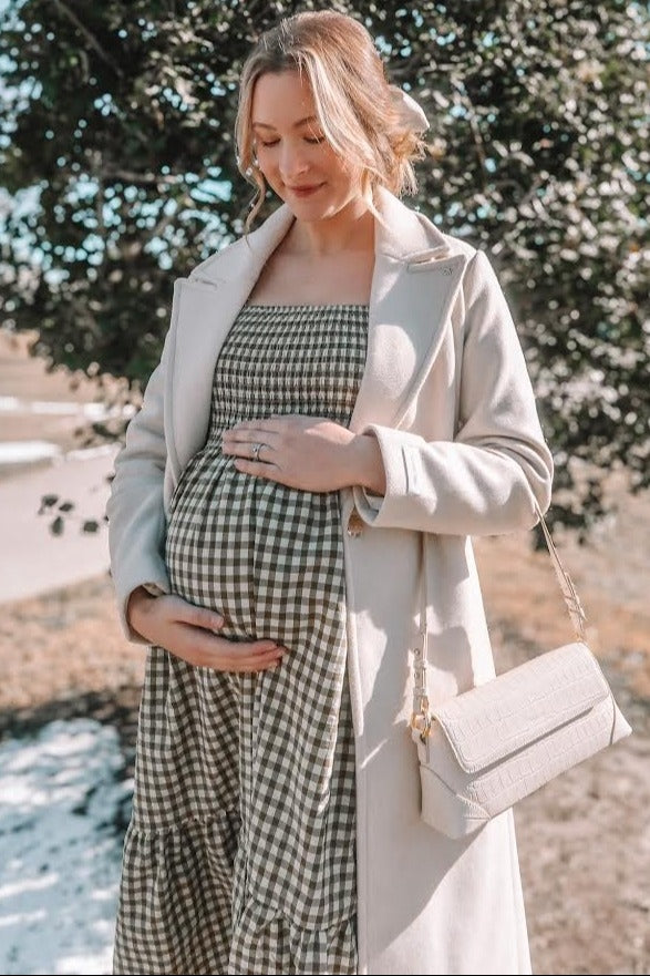 woman wearing a green and white checked maternity dress with a beige coat, cradling her baby bump