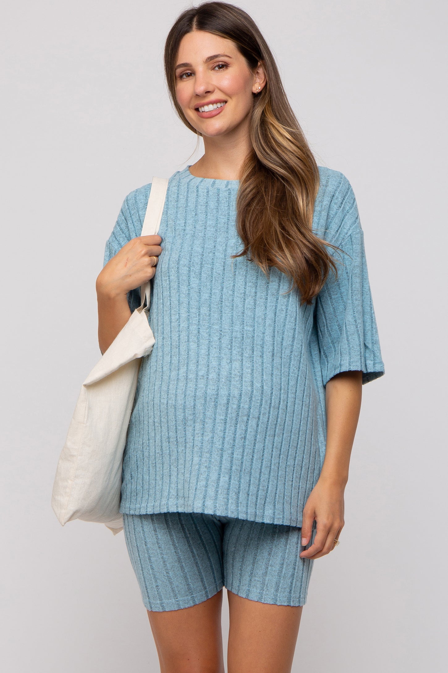 woman wearing a light blue ribbed maternity top and matching shorts, holding a tote bag and showing her baby bump