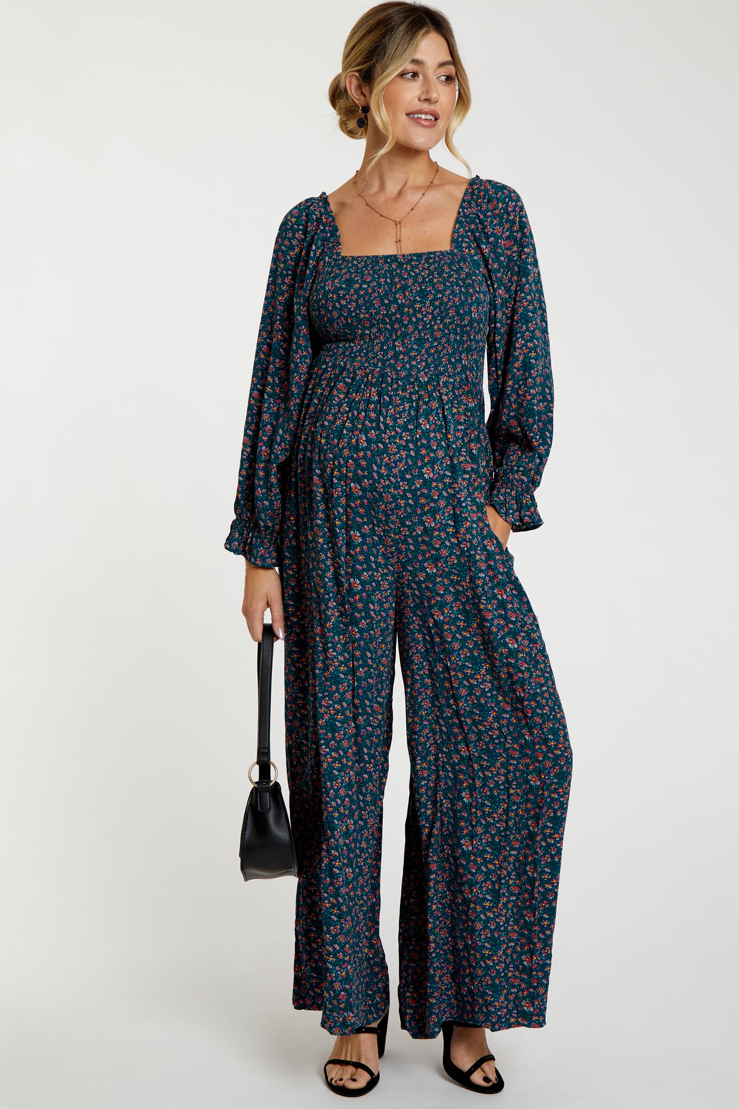 woman wearing a dark floral maternity jumpsuit with a square neckline and long sleeves, holding a black handbag