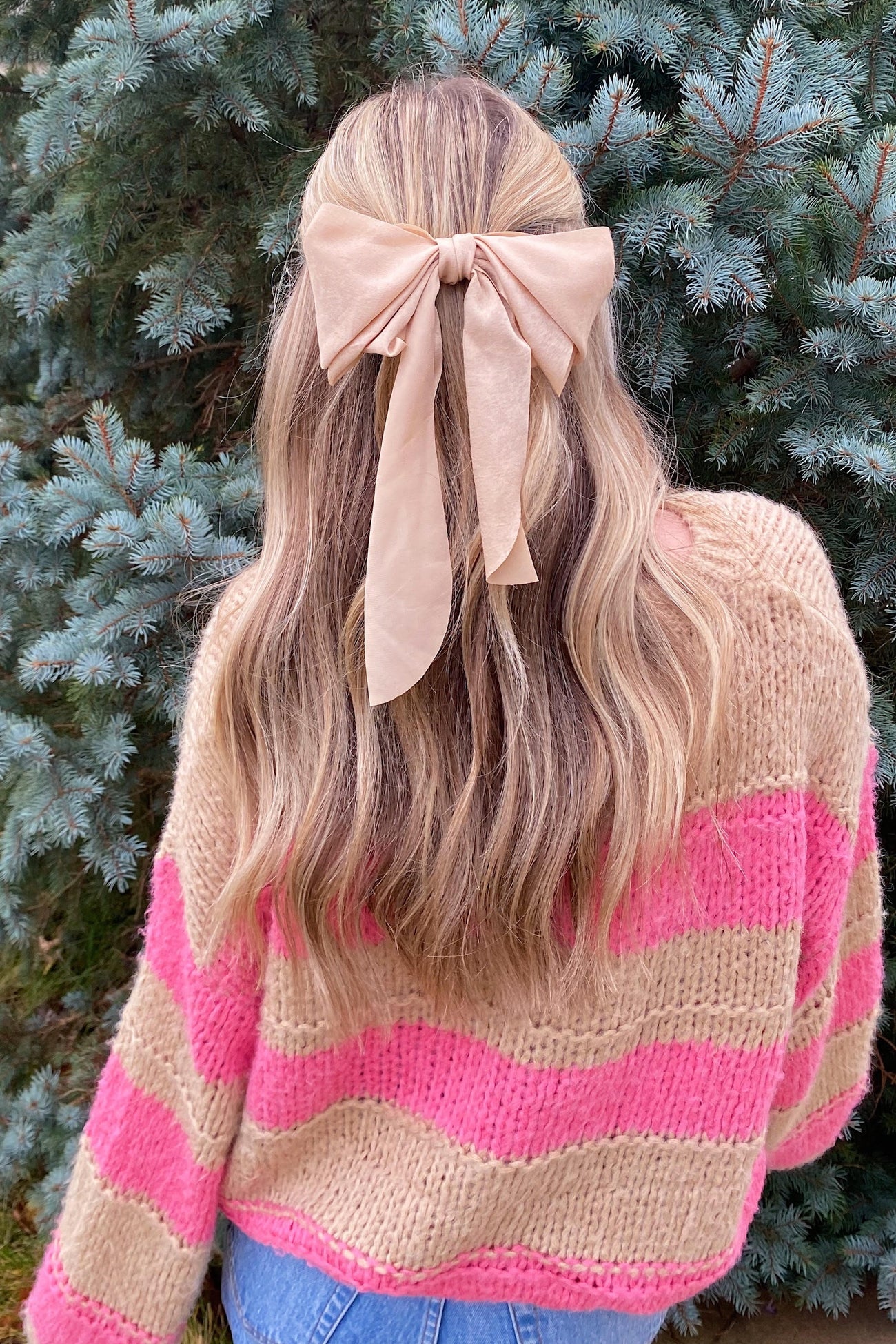 woman wearing a pink and beige striped sweater with a large beige bow in her hair, standing in front of evergreen trees