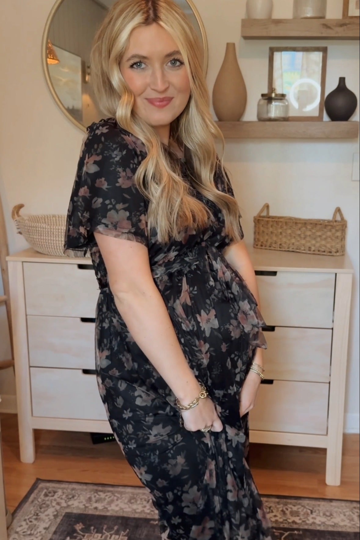 woman wearing a black floral maternity dress, standing with her baby bump visible in a stylish home setting